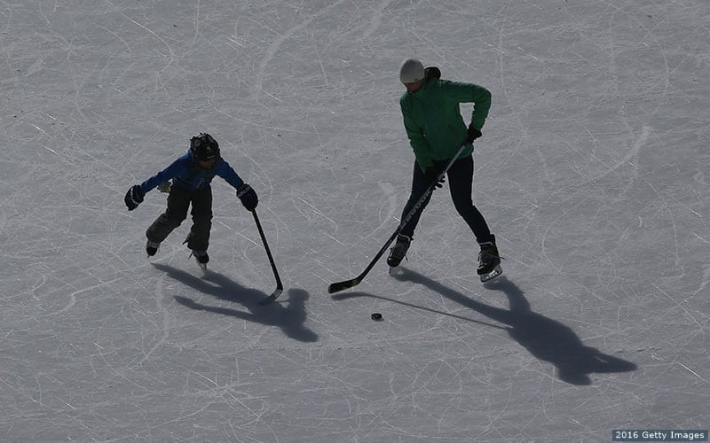 A mother and her son practice on an ice pad adjacent to outdoor shinny hockey action on a lake.