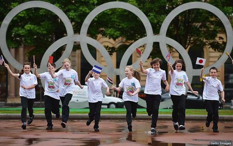 Children with flags from around the world play together in front of the Olympic rings.
