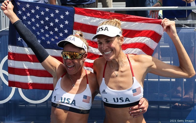 U.S. beach volleyball athletes April Ross (left) and Alix Klineman hold the American flag aloft in celebration after winning gold at the 2020 Olympic Games.