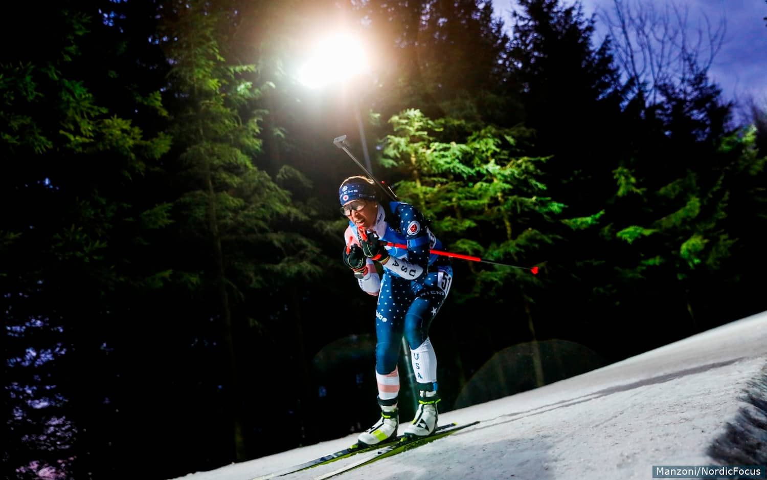 Susan Dunklee skis under the lights at night and in front of a row of evergreens with her poles tucked under her arms.