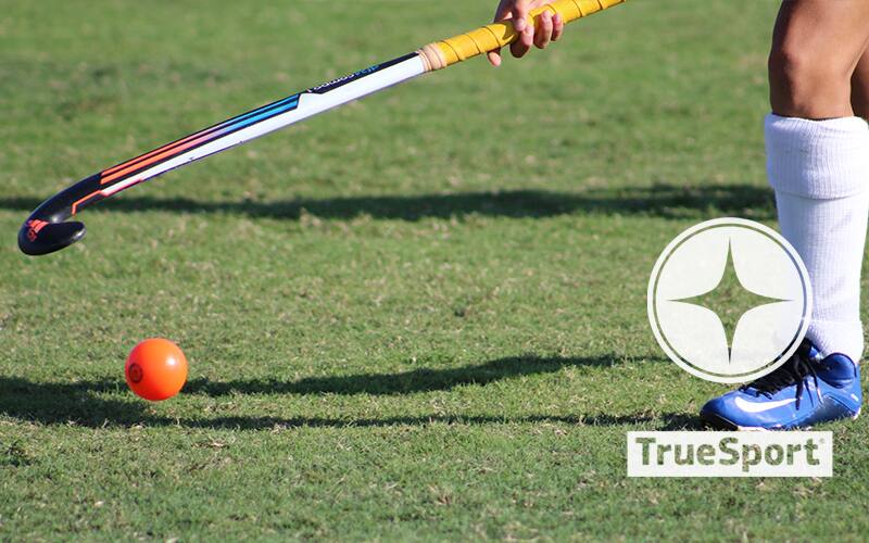 Field hockey stick and ball on grass with TrueSport logo