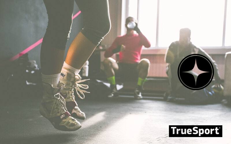 Woman in training gym jumping rope with men sitting behind her, one of them is drinking water