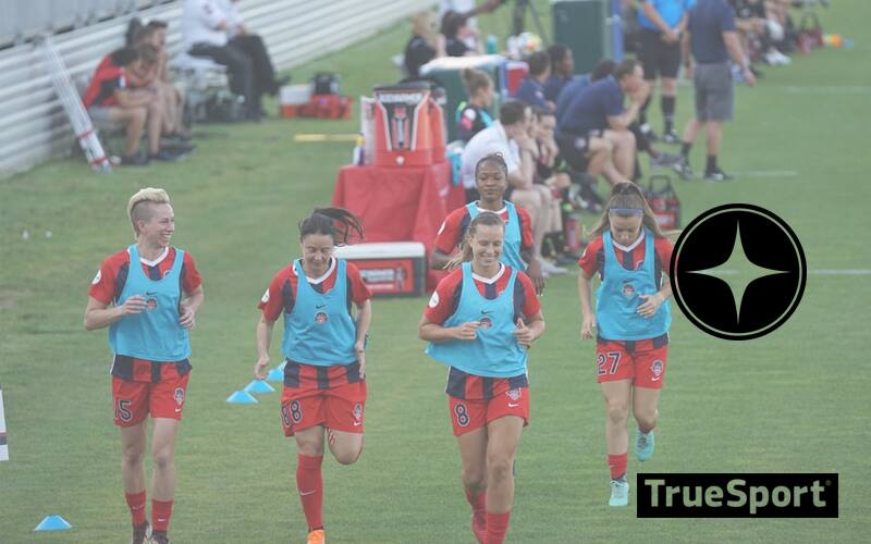 Athletes running on field, all are women wearing red shorts and socks with a light blue top
