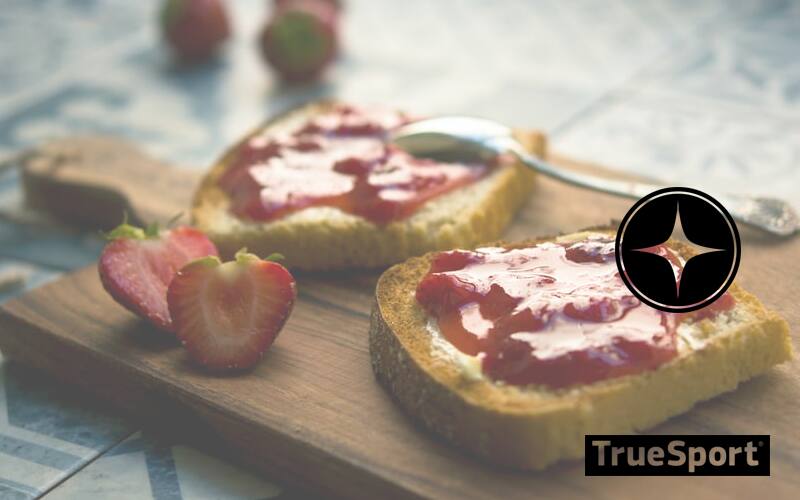 Two pieces of bread on a wooden board coated in a red jelly, two strawberry halves sit on the side of the board