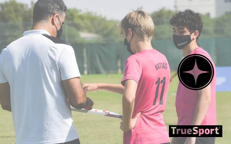 Coach wearing a white polo and black masks is talking with two younger male athletes wearing pink jerseys 