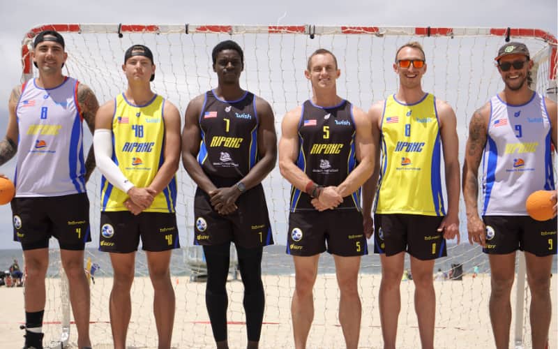 Men's Beach Team athletes standing in front of a beach handball goal, shoulder to shoulder