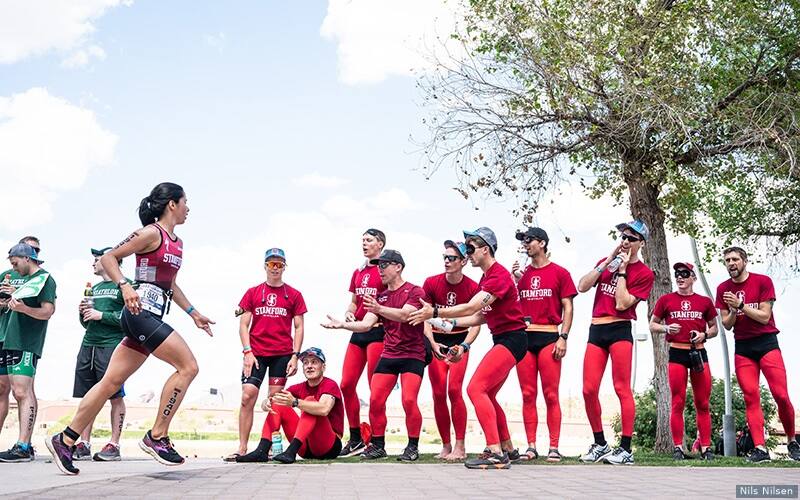 triathletes on the University of Stanford club triathlon team dressed in red tights give a woman Stanford triathlete high fives during a race.
