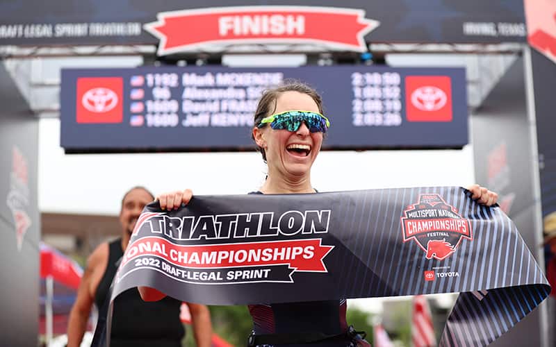 woman with brown hair wearing sunglasses and smiling while holding the national championships banner