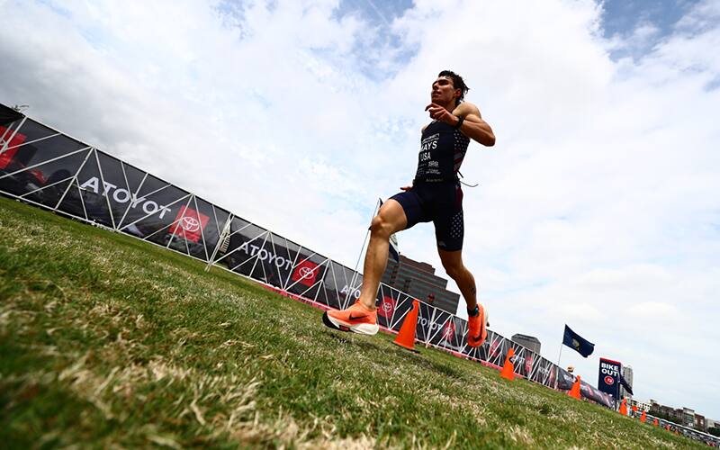 Photograph of 18 year old man wearing a dark blue Team USA triathlon kit and orange shoes running on green grass through the transition area at a triathlon