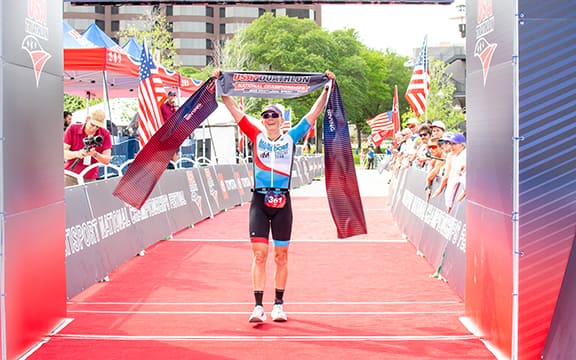female triathlete stands at finish line with finish line tape above head