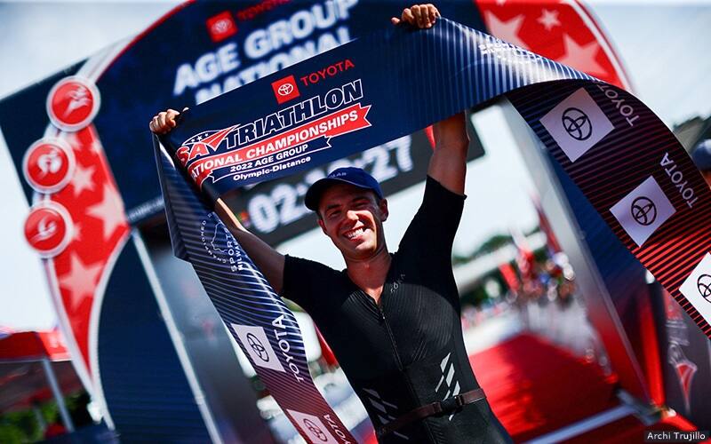 male triathlete holds the race finish banner over his head 