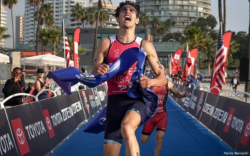 male triathlete runs across the finish line, holding the finisher's tape. he has an excited expression on his face.