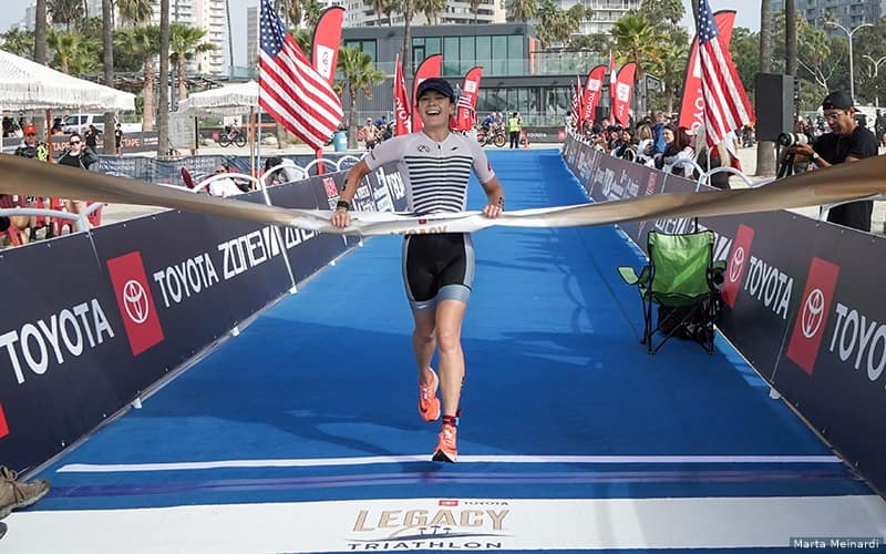 Female triathlete Jenna Haufler runs across the finish line, grabbing the finisher's tape. She is smiling and wearing a white jersey with black stripes.
