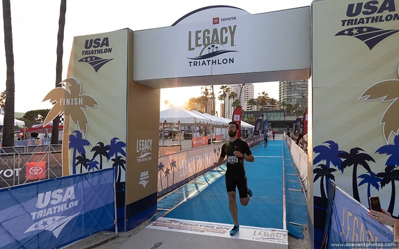 man running through the finish line of the Toyota Legacy Triathlon. he is running on blue carpet underneath a gold and white finish line arch.