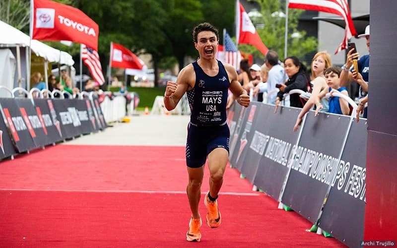 A young triathlete wearing a blue triathlon suit runs toward the finish line with an excited expression on his face