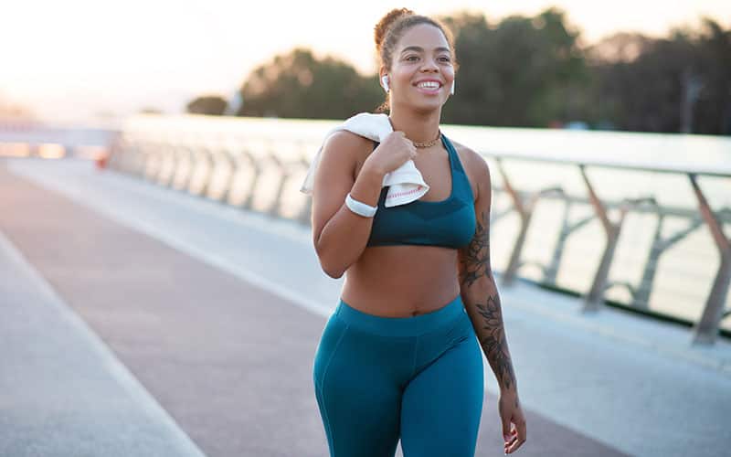 A woman in workout clothing walks while smiling and holding a towel over her shoulder