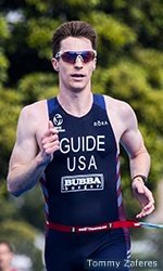 photo of zach goodman running toward the finish line wearing a dark blue triathlon racing kit with Guide written across his chest